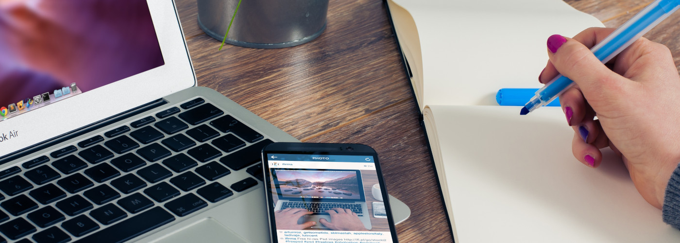 Ein Laptop und ein Smartphone liegen auf einem Tisch. Eine Hand hält einen blauen Stift und schreibt in ein offenes Notizbuch.