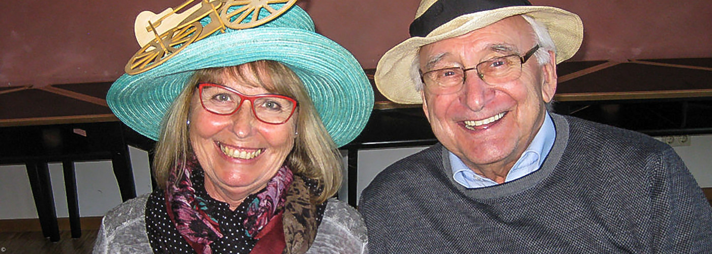 Zwei Senioren nebeneinander, die Frau links trägt einen türkisfarbenen Hut, der Mann rechts einen hellen Strohhut.