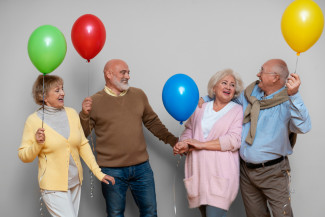 Vier ältere Personen stehen mit bunten Luftballons in den Händen, tragen lässige Kleidung in gedeckten Farben.