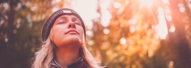 Eine Frau mit langem, blondem Haar und schwarzer Strickmütze vor herbstlichem Hintergrund mit Sonnenlicht