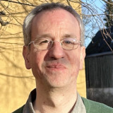 herr Vierling mit grauen Haaren und Brille vor einer gelben Hauswand und einem dunklen Holzzaun im Freien