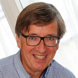 Mann mit kurzem braunem Haar trägt hellblaues Hemd mit dunklen Punkten, sitzt vor hellem Fenster.