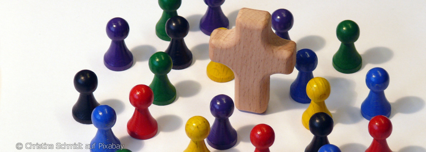 Holzkreuz umgeben von bunten Spielfiguren auf weißer Fläche